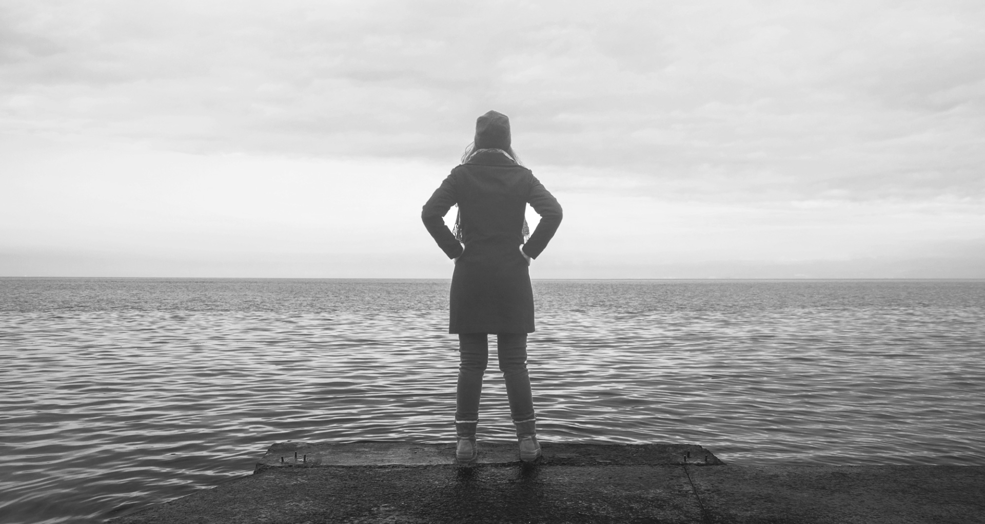 A woman wearing a hat, boots, and a winter coat stands at the edge of a lake with her hands on her hips.