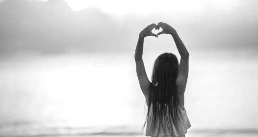 A girl with long hair holds her arms above her head, making a heart shape with her hands.