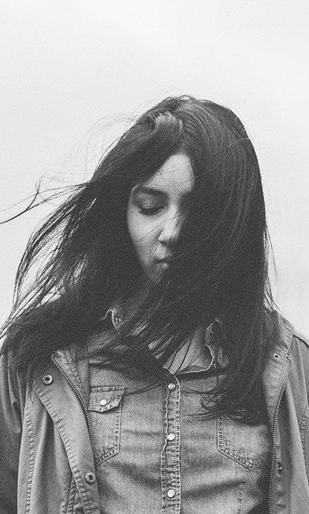 A woman wears a denim shirt and jacket while the wind blows her dark hair across her face.