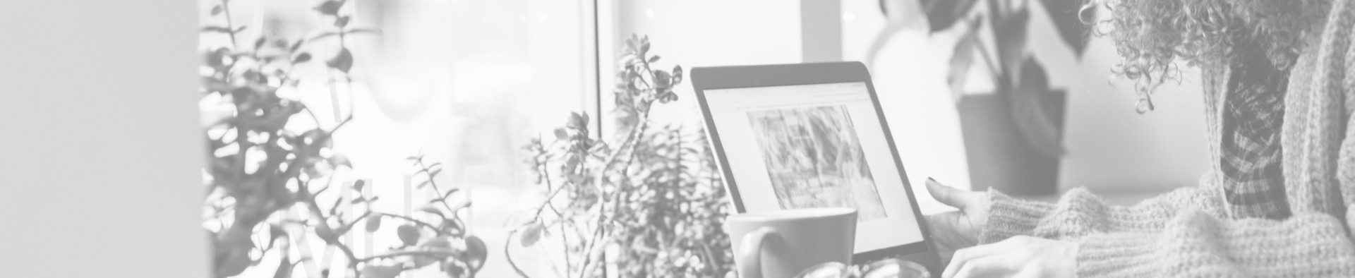 A woman wearing a sweater looks at an image on her laptop, which is sitting on a desk with a coffee mug and plants next to it.