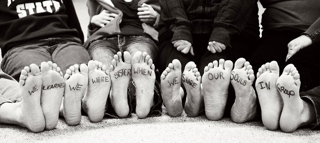 Six people's feet are lined up with words written on them, reading "We learned we were sisters when we shared our souls in group."