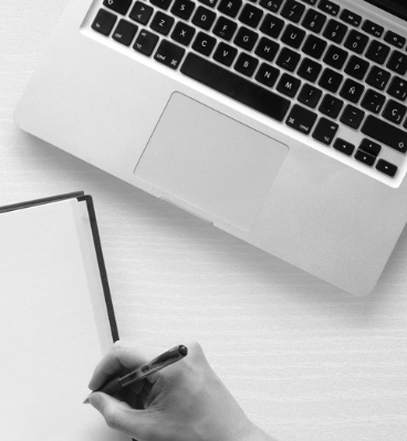 A hand holds a pen, ready to write on a blank sheet of notebook paper, with a laptop off to the side.