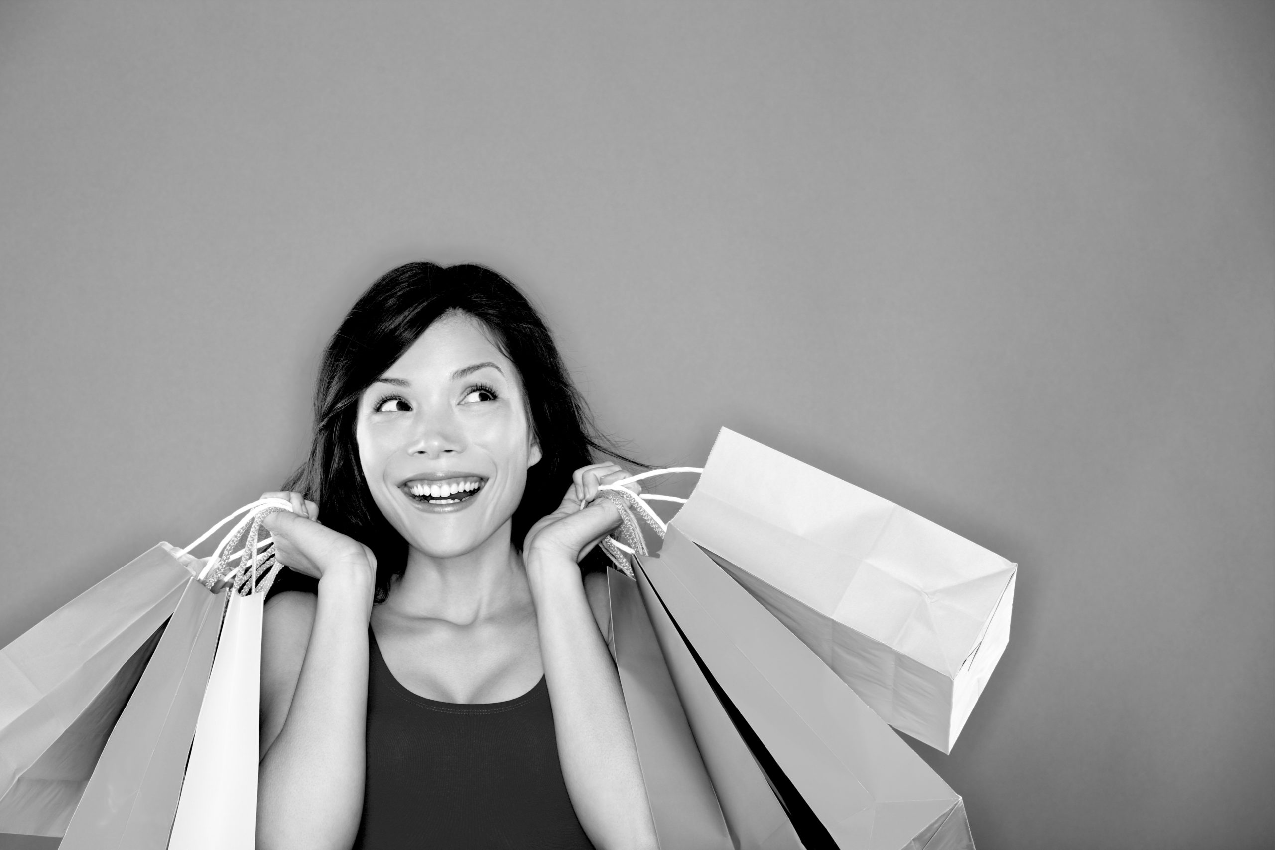 A smiling woman wearing a black tank top holds paper shopping bags in both hands.